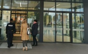 Na zdjęciu widoczny umundurowany policjant, w obecności dwóch kobiet. W tle drzwi wejściowe do punktu usługowo handlowego.