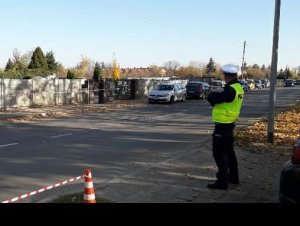 Na zdjęciu widoczny umundurowany policjant w trakcie akcji Znicz. Zdjęcie wykonane na tle cmentarza.