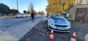 Na zdjęciu widoczny umundurowany policjant w trakcie akcji Znicz. Zdjęcie wykonane na tle cmentarza.