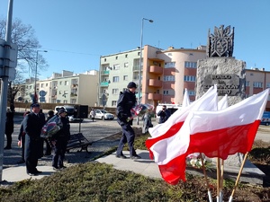 Na zdjęciu widoczne umundurowane policjantki w asystencie strażaka (mundur galowy) , które składają kwiaty pod pomnikiem ZESŁAŃCÓW SYBIRU. Zdjęcie wykonane na zewnątrz w tle flagi Polski.