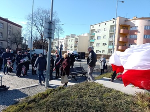 Na zdjęciu widoczne umundurowane policjantki w asystencie strażaka (mundur galowy) , które składają kwiaty pod pomnikiem ZESŁAŃCÓW SYBIRU. Zdjęcie wykonane na zewnątrz w tle flagi Polski.