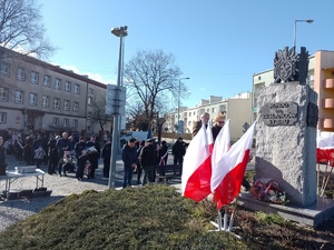Na zdjęciu widoczne umundurowane policjantki w asystencie strażaka (mundur galowy) , które składają kwiaty pod pomnikiem ZESŁAŃCÓW SYBIRU. Zdjęcie wykonane na zewnątrz w tle flagi Polski.