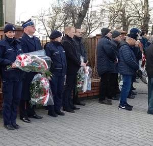 Na zdjęciu widoczne umundurowane policjantki w asystencie strażaka (mundur galowy) , które składają kwiaty pod pomnikiem ZESŁAŃCÓW SYBIRU. Zdjęcie wykonane na zewnątrz w tle flagi Polski.