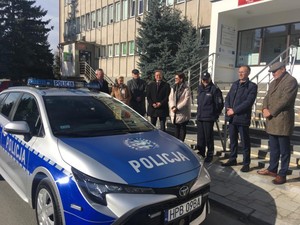 Na zdjęciu widoczny umundurowany policjant Komendant Powiatowy Policji w Strzelinie w obecności przedstawicieli samorządu lokalnego. Za zgromadzonymi osobami widoczny radiowóz oznakowany. Zdjęcie wykonane na zewnątrz na tle budynku Starostwa Powiatowego w Strzelinie