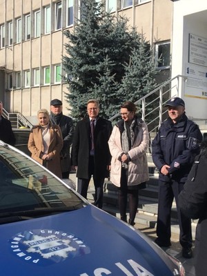 Na zdjęciu widoczny umundurowany policjant Komendant Powiatowy Policji w Strzelinie w obecności przedstawicieli samorządu lokalnego. Za zgromadzonymi osobami widoczny radiowóz oznakowany. Zdjęcie wykonane na zewnątrz na tle budynku Starostwa Powiatowego w Strzelinie