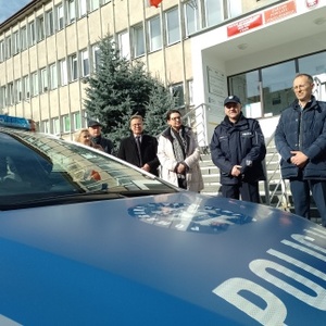 Na zdjęciu widoczny umundurowany policjant Komendant Powiatowy Policji w Strzelinie w obecności przedstawicieli samorządu lokalnego. Za zgromadzonymi osobami widoczny radiowóz oznakowany. Zdjęcie wykonane na zewnątrz na tle budynku Starostwa Powiatowego w Strzelinie