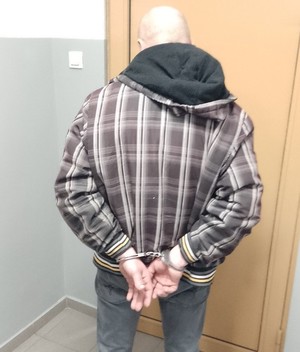 Na zdjęciu widoczny zatrzymany mężczyzna, który posiada założone kajdanki na ręce trzymane z tyłu. Zdjęcie wykonane w pomieszczeniu. Zatrzymany mężczyzna stoi na tle zamkniętych drzwi.