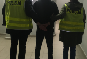 Na zdjęciu widoczni nieumundurowani policjanci z kamizelkami odblaskowymi z napisem POLICJA. Pomiędzy policjantami stoi mężczyzna, który posiada założone kajdanki na ręce trzymane z tyłu. Zdjęcie wykonane w pomieszczeniu zamkniętym.