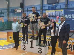 Na zdjęciu widoczni uczestnicy zawodów, którzy stoją na podium ponumerowanym od 1 do 3 miejsca. Zdjęcie wykonane na hali sportowej. Tuż obok uczestników stoją organizatorzy.
