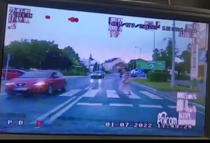 Na zdjęciu widoczne przejście dla pieszych, na którym znajduje się piesza oraz przejeżdżający samochód osobowy koloru ciemnego