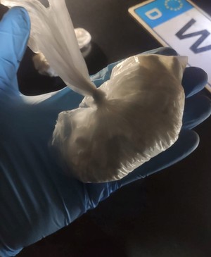 Na zdjęciu widoczny plastikowa reklamówka wraz z zawartością w postaci środków odurzających (białe owalne tabletki). W tle widoczny fragment tablicy rejestracyjnej.