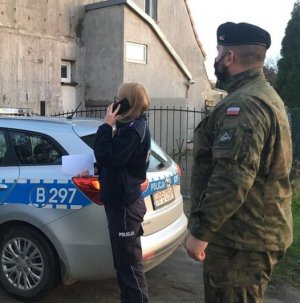 Zdjęcie zawiera wizerunek policjantki oraz żołnierza Wojsk Obrony Terytorialnej WOT podczas kontroli osób objętych kwarantanną domową na tle radiowozu oznakowanego oraz budynku mieszkalnego.