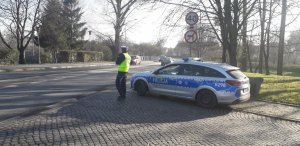 Fotografia zawiera wizerunek policjanta drogówki w trakcie wykonywania czynności służbowych w ramach akcji &quot;Trzeźwy poranek&quot;. Policjant na tle radiowozu oznakowanego przy drodze wojewódzkiej w obrębie terenu zabudowanego.