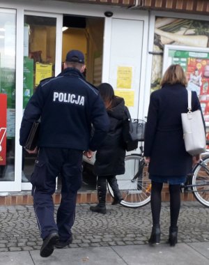 Fotografia zawiera sylwetkę umundurowanego funkcjonariusza Policji, który wchodzi na teren lokalu usługowo - handlowego celem dokonania kontroli. Z prawej strony funkcjonariusza dwie kobiety, które są przedstawicielkami strzelińskiego Sanepidu.