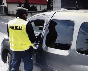 Na pierwszym palnie fotografii funkcjonariusz umundurowany w czapce z kamizelką odblaskową z napisem POLICJA podczas kontroli kierującego samochodem osobowym koloru ciemno szarego. W tle budynki mieszkalne.