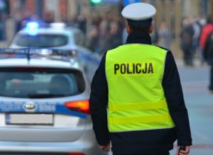 Zdjęcia zawiera umundurowanego policjanta Ogniwa Ruchu Drogowego, funkcjonariusz posiada granatowy mundur, kamizelkę odblaskową oraz czapkę koloru białego. Z lewej strony zdjęcia radiowóz oznakowany. W tle zdjęcia (drugi plan) zatłoczona ulica.