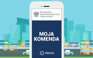 Na ikonografii widoczny telefon komórkowy w kolorze biały, na ekranie którego znajduje się zainstalowana aplikacja mobilna Moja Komenda. W tle budynki, ludzie poruszający się na chodniku oraz samochodu osobowe.