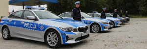 Na zdjęciu widoczne oznakowane radiowozy marki BMW ustawione równolegle do ulicy. Tuż obok radiowozów stoją umundurowaniu policjanci Ruchu Drogowego.