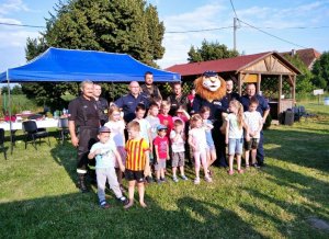 Na zdjęciu widoczne dzieci w wieku szkolnym biorące udziała w zajęciach profilaktycznych odbywających się na terenie placu zabaw. W tle widoczni umundurowani policjanci oraz maskotka Komisarz Lew.