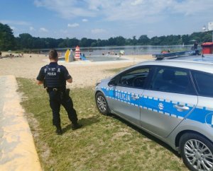 Na zdjęciu widoczny umundurowany policjant w trakcie kontroli akwenu wodnego. W tle osoby w strojach kąpielowych korzystające z atrakcji.